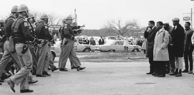 March 7. Hosea Williams (at right front in dark raincoat) and John Lewis (at right in light-colored coat), led protesters to the Edmund Petis bridge on their way from Selma to Montgomery, Alabama during the Civil Rights Movement. Officers are seen giving the protestors a 2 minute warning to turn around before they tear gassed and beat the marchers. Credit: Photo taken by Spider Martin. National Archives photo.