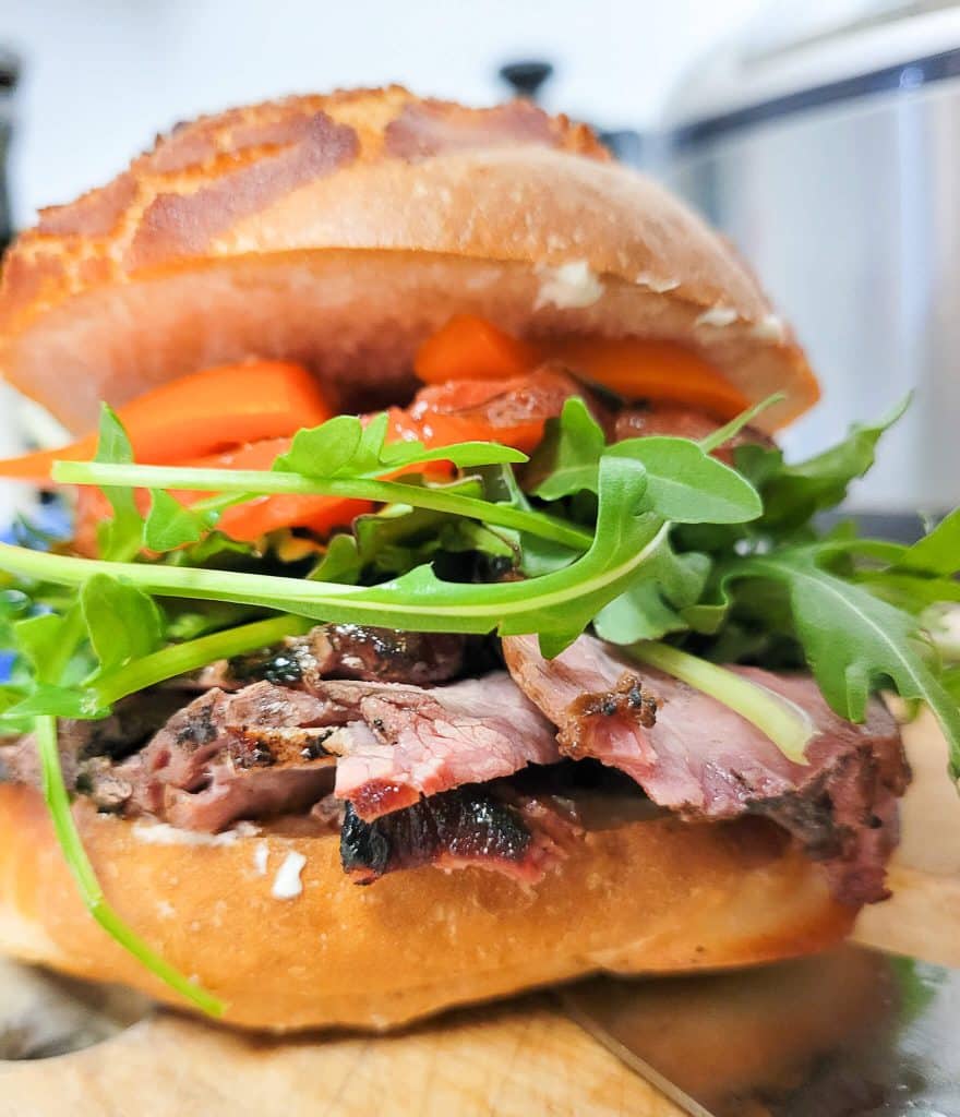 A smoked tri tip sandwich on dutch crunch roll with arugula, cilantro, smoked tomato, bell pepper, and horseradish sauce.