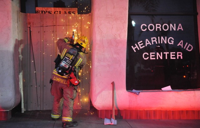 A Corona firefighter cuts through a metal door as firefighters battled a blaze at the Corona Mall Tuesday. There were no injuries. A total of 31 firefighters responded, containing the fire to a single unit. Credit: Photo by Jerry Soifer