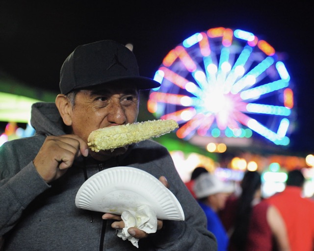 Featured Photos 10-18-2024. Corona resident Hector Manriquez enjoys corn on the cob as the Ferris wheel turns behind him at the St. Edward Catholic Church Carnival in Corona. Credit: Photo by Jerry Soifer