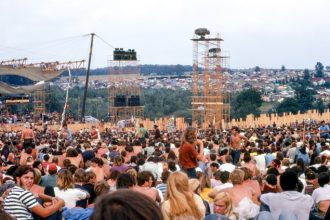 August 15. Joe Cocker performs on stage at left before crowd and huge lighting/sound towers. Cars and tents can be seen in the background. Credit: Woodstock Whisperer