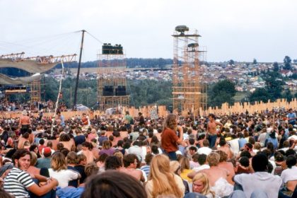 August 15. Joe Cocker performs on stage at left before crowd and huge lighting/sound towers. Cars and tents can be seen in the background. Credit: Woodstock Whisperer
