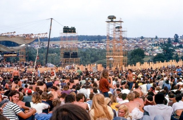 August 15. Joe Cocker performs on stage at left before crowd and huge lighting/sound towers. Cars and tents can be seen in the background. Credit: Woodstock Whisperer